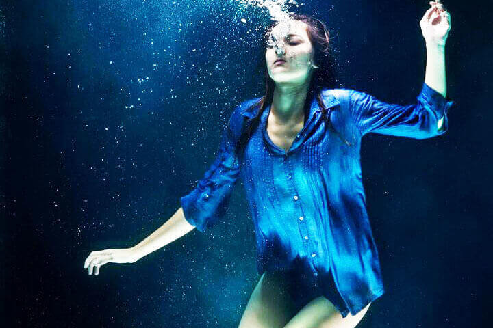 Foto de uma mulher com camisa azul, mergulhada em uma piscina, de olho fechado, soltando bolinhas de ar pela boca como se estivesse afundando e se afogando. A foto ilustra o artigo Como a Psicanálise trata a depressão escrito no blog do psicanalista Homero Mônaco para o site A Sessão de Terapia.