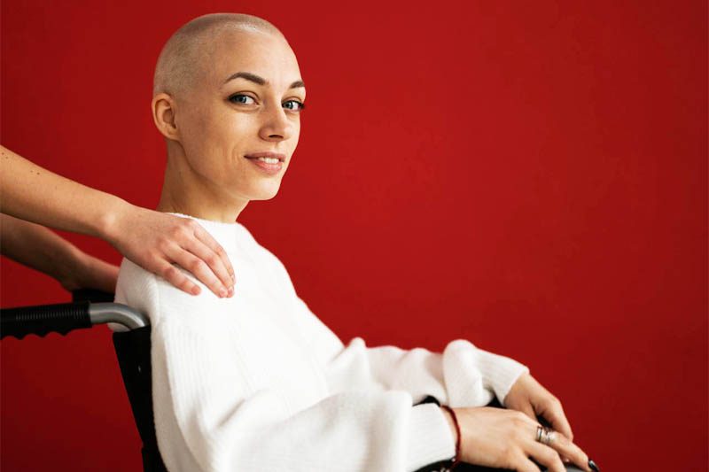 Foto de uma mulher branca, com o cabelo raspado, sentado em uma cadeira de rodas que está sendo empurrada por alguém. A mulher olha sorrindo para a câmera como se tivesse passado por uma cura e uma recuperação emocional. A foto ilustra o artigo O Poder da Terapia para a sua Recuperação Emocional escrito pelo psicanalista Homero Mônaco para o site Online Psicanalista.