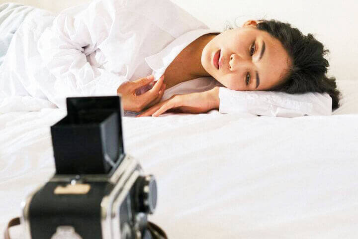 Foto de uma mulher deitada de lado em sua cama, e à sua frente está uma câmera. Ela está com cara de pensativa tentando entender as suas emoções. A foto ilustra o artigo 7 Passos para Resolver Conflitos Internos de Relacionamento escrito no blog do psicanalista Homero Mônaco para ao site A Sessão de Terapia.