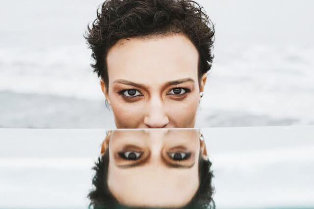 Foto de uma mulher olhando para a câmera enquanto o seu rosto se reflete em um espelho abaixo do seu rosto. A foto ilustra o artigo Reconstruindo a sua Identidade Através da Aceitação e da Transformação escrito pelo psicanalista Homero Mônaco para o site Online Psicanalista.