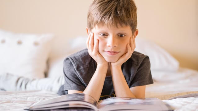 Foto de um menino deitado de bruços na cama, com a cabeça levantada apoiada pelas duas mãos e com um livro . A foto ilustra o artigo Como curar Traumas com a Psicanálise escrito pelo psicanalista Homero Mônaco para o site Online Psicanalista.
