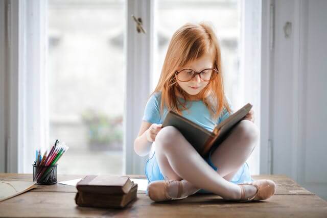 Foto de uma menina ruiva, de óculos, sentada em cima de uma mesa com um livro nas mãos. Ela parece feliz como se estivesse fazendo uma leitura por prazer. A foto ilustra o artigo Leitura por prazer: Novo estudo revela benefícios surpreendentes sobre ela escrito pelo psicanalista Homero Mônaco para o site Online Psicanalista.