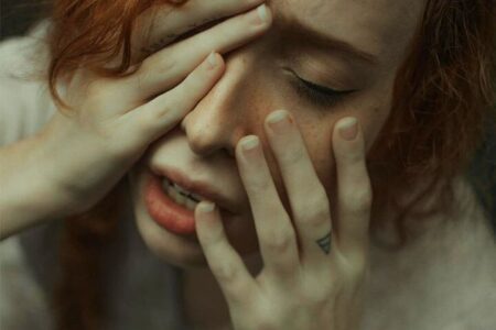 Foto do rosto de uma mulher ruiva, com expressão desesperada e parecendo estar com ansiedade e com as mãos em seu rosto. A foto ilustra a página do Perfil do Psicanalista Homero Mônaco para o site A Sessão de Terapia.