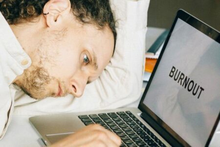 Foto de um homem com cabeça deitada em seu braço esquerdo, que está apoiado em uma mesa, olhando para o seu notebook que na tela está escrito Burnout. A foto ilustra a página do Perfil do Psicanalista Homero Mônaco para o site A Sessão de Terapia.