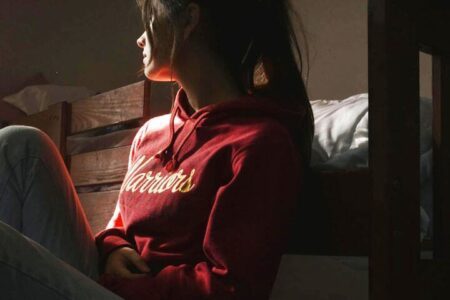Foto de uma jovem de lado, sentada no chão com as costas apoiadas em sua cama, olhando para o nada. O ambiente está escuro e ela parece estar com uma crise de depressão. A foto ilustra a página do Perfil do Psicanalista Homero Mônaco para o site A Sessão de Terapia.