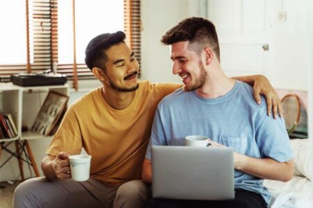 Foto de um jovem oriental, com uma caneca branca na mão esquerda, sentado em um sofá, abraçando com o braço esquerda um outro rapaz, também sentado no mesmo sofá, com um notebook em seu colo. Ambos estão sorrindo um para o outro e representando os seus gêneros. A foto ilustra a página do Perfil do Psicanalista Homero Mônaco para o site A Sessão de Terapia.
